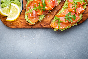 Avocado sandwich with salmon on rye bread with guacamole sauce, arugula and sesame seeds on wooden board on gray table background, top view