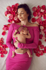 beautiful family portrait mum in a pink dress cuddles her newborn daughter lying on the bed by the window. baby is lying on its mother's stomach with pink rose petals.