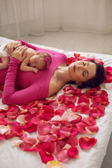 beautiful family portrait mum in a pink dress cuddles her newborn daughter lying on the bed by the window. baby is lying on its mother's stomach with pink rose petals.