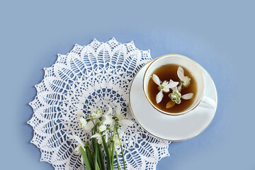 Spring tea party: a cup of tea, a bouquet of snowdrops on an openwork napkin, blue background - obrazy, fototapety, plakaty