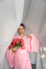 Beautiful portrait brunette woman in a pink Asian dressing gown standing in front of the window against the curtains with red tulips for a holiday.