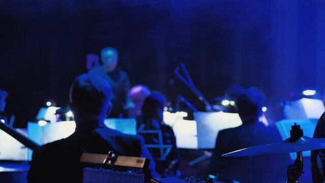 Part of an orchestra during a performance or rehearsal on stage with the conductor. Back view shows the backs of the musicians and instruments under dramatic stage lightingju Blurred video