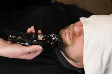 The clients head is covered with a towel during a beard trim. Shortening the length of the beard from the sides by the master for the client.