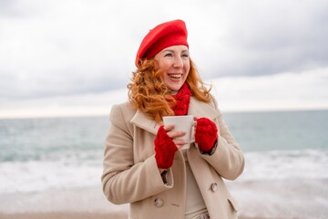 Obraz premium Woman beach in red beret scarf and mitts holding a cup of tea in his hands. Depicting beach relaxation and cozy attire. Walks by the sea
