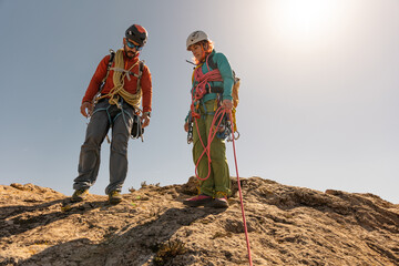 Two people are standing on a rocky hill, one of them wearing a red jacket. They are both wearing...