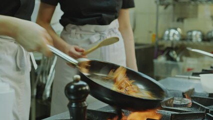 Head chef teaching young female apprentice how to flip food in skillet and cook above fire during culinary internship in restaurant kitchen