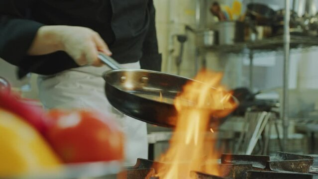Restaurant chef in apron flipping food ingredients in cooking pan over fire flames above stove in the kitchen. Midsection, close-up shot