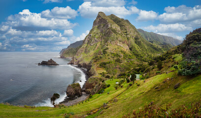 The photo depicts the picturesque Madeira: green hills, rocky cliffs, blue sea, and white clouds....