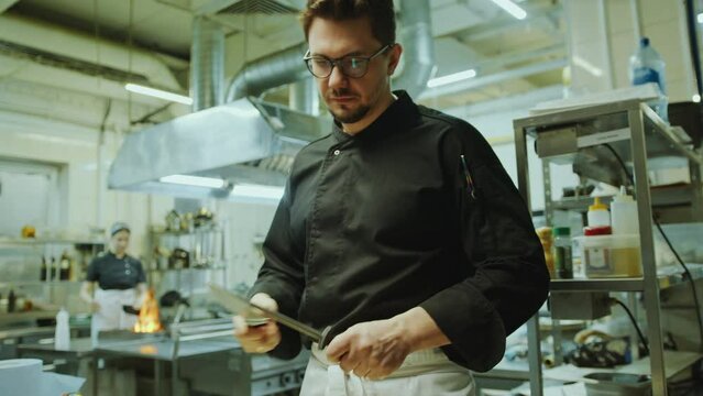 Professional chef standing in restaurant kitchen and sharpening knife with honing steel before cooking food