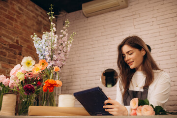 Portrait of attractive woman florist working on tablet at workplace. Concentrated female sitting at counter in floral store and typing on digital tablet.