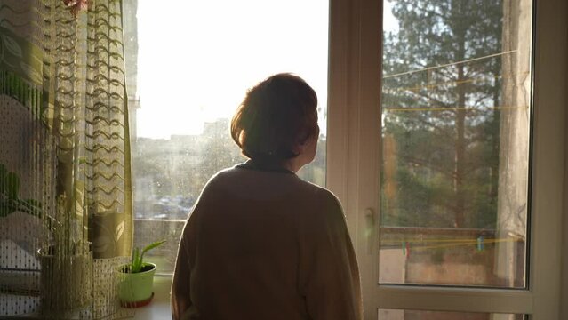 Back view of elderly woman walking in the old soviet apartment to watch through the window. Sunset light comes to the room, dirty windows. Lonely granny waiting for her family.