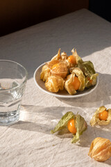 Ripe physalis berries in shells, glass with water on neutral beige tabletop, linen tablecloth,...