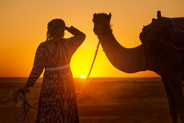 Silhouette of woman in traditional national clothes holding camel by rope while admiring sunset in desert - obrazy, fototapety, plakaty