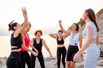 Fototapeta na wymiar Group of adult athletic multiethnic women in sportswear happily jumps at coast in background of sunset and sea. Concept of wellness and female friendship