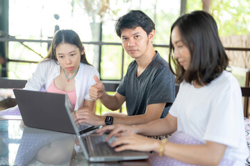 Asian male students give a thumbs up like symbol and three female classmates sit and type on a project on a laptop in a university faculty library, wearing casual clothes and giving confident glances.