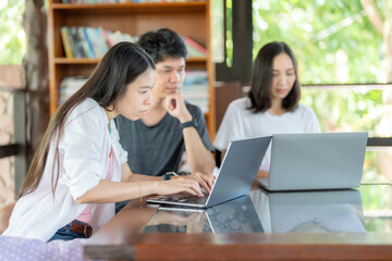 Side view of an Asian female student studying online on a laptop and two friends in the library. They are studying in a university group and read books every week to gain knowledge.