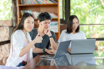 Side view of Asian female student with 2 friends studying online on laptop, thumbs up, like sign at university library, group reading books every week to gain knowledge and intelligence.