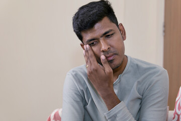 indian young man sadly sitting on the sofa at home and looking at the camera and wiping his tears