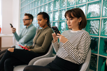 Serious multicultural young business people sitting in row using smartphones, waiting for job interview, sitting in queue line row in chairs in modern office waiting room.