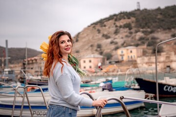 Fototapeta na wymiar Woman holds yellow tulips in harbor with boats docked in the background., overcast day, yellow sweater, mountains