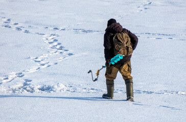 A man fisherman with an ice auger in his hands leads through the white snow in winter - 768591434