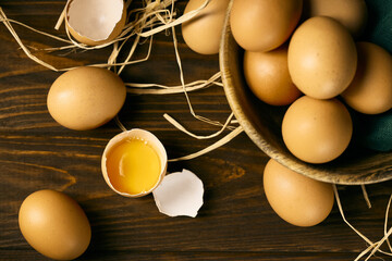 Raw eggs on a brown woody background  with one broke egg on a straw pillow, top view, brown bowl with eggs inside