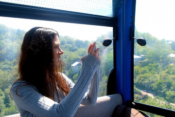 Woman tourist recording video on smartphone sitting in funicular of cable car in mountains. Pretty happy brunette female exploring world travelling in Georgia, Batumi. Travel, tourist, wanderlust. - obrazy, fototapety, plakaty