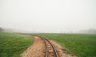 Narrow gauge railway in the foggy green field. The track curves away from the viewer to the left,...
