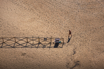 Aerial view of a man walking along the beach towards his towel, leaning on a wooden railing
