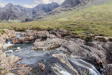 A clear stream cascades through weathered rocks at the Fairy Pools, set against the dramatic...