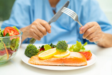 Asian elderly woman patient eating Salmon steak breakfast with vegetable healthy food while sitting and hungry on bed in hospital.