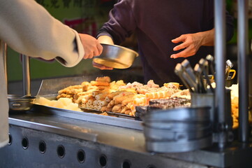TW-03.03.24: At an oden stall, owner hands containers to customers. Taiwan's oden, influenced by Japan, is a popular stew with ingredients like daikon, konjac, fish cakes, simmered in flavorful broth.