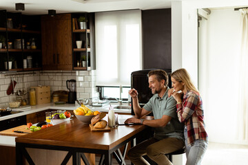 Cheerful couple enjoys a light-hearted moment in their sunny kitchen, working on laptop surrounded by a healthy breakfast - 768598055