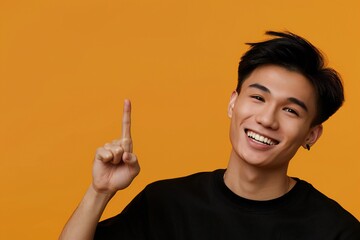 asian american male person with toothy smile on face pointing with index finger on empty space over orange background. Handsome young guy indicating place for advertising text in studio