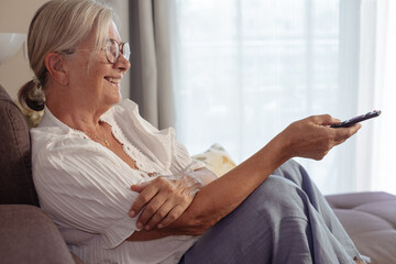 Happy carefree senior white haired woman sitting on home sofa holding tv remote control. Elderly relaxed lady watching television, movie or social entertainment