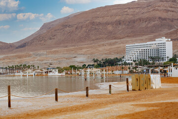 Beach resort near a mountain. The sandy shores show wooden posts connected by ropes. Palm trees and a large white building are visible, with mountain rocky hills and sea. Morning sky, rows umbrellas