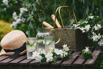 fresh lemonade with mint in summer garden. Blooming Philadelphus coronarius (Sweet Mock-orange, Jasmine) bouquet on wooden table with gardener tools. Summertime relax