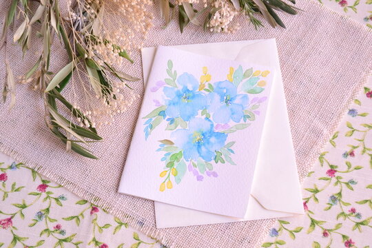 Wedding invitation blue watercolor hand painted flowers white envelope on burlap and olive leaves background, romantic style party ceremony, artisan greeting card painting, small home craft business