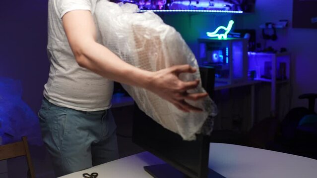 Cropped shot of unrecognizable young man packing desktop monitor in cardboard box and bubble wrap in dark room during preparation to relocation new home. Relocating delivery service concept.