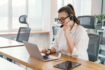 A concentrated businesswoman with a headset engages in a discussion on her laptop in a well-lit,...