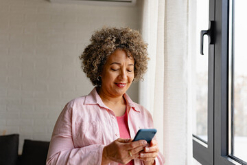 A mature woman of mixed race standing near large window, holding a smartphone and smiling. Mobile phone apps for older people - 768640868