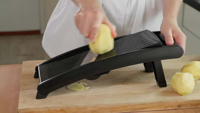 Chef grating potatoes into slices on a mandolin grater, preparing healthy food, wooden table, home cooking, vegetarian food, vegetables rich in vitamins, diet. Close-up, front view