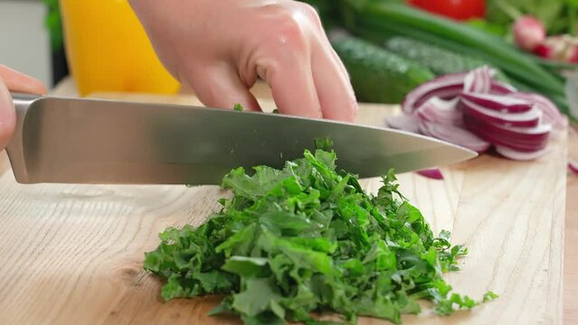 The chef cuts greens, young kale leaves, parsley, and cilantro on a wooden board with a sharp knife. Healthy eating concept. Close-up, side view, vegetables in the background