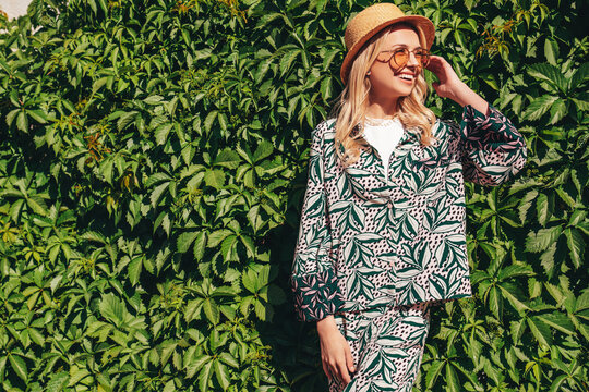 Young beautiful smiling blond hipster woman in trendy summer hippie costume clothes. Carefree female posing in street at sunny day. Positive model outdoors. Cheerful and happy in hat near green bushes