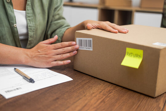 A female shopper affixes a Return sticker, featuring a yellow label with a barcode, onto a cardboard box, initiating the online shopping refund process. 