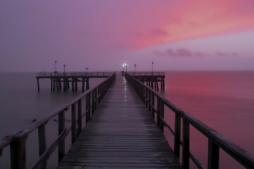 Obraz premium A tranquil pier extends into a calm sea under a captivating pink and purple sky, illuminated by distant lights