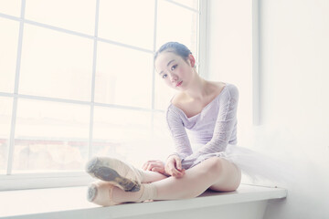 young Japanese ballerina in a tutu and pointe shoes poses in a photo studio sitting on a windowsill