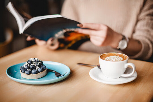 A serene scene unfolds as a woman indulges in a book, accompanied by a delightful blueberry tart and a steaming cup of coffee, creating a warm and inviting ambiance.
