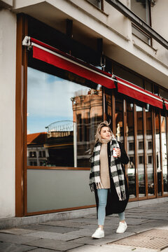 A stylish woman in a black coat and plaid scarf walks gracefully along a city sidewalk, the vibrant red awning adding a pop of color to the urban scene.