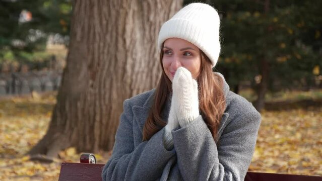 Attractive woman in stylish grey coat, warm white hat and mittens enjoying autumn day on bench in park